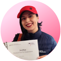 Teilnehmerin mit roter Mütze hält ein Zertifikat „Deutsch-Test für Zuwanderer“